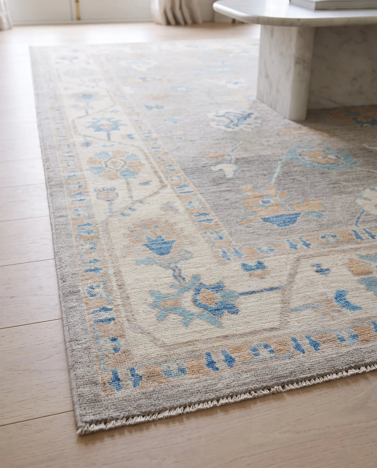 Hand-knotted Afghan wool rug with a muted grey, blue, and beige floral Oushak pattern on a light timber floor.