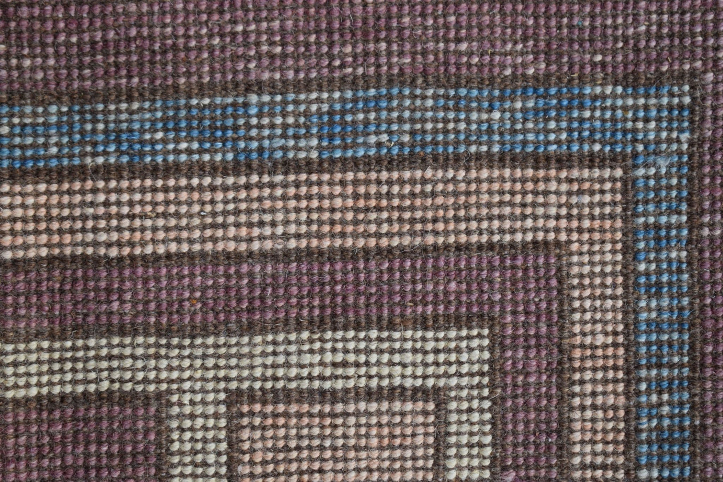 A close-up of a handmade wool rug featuring geometric borders in purple, blue, and cream tones.