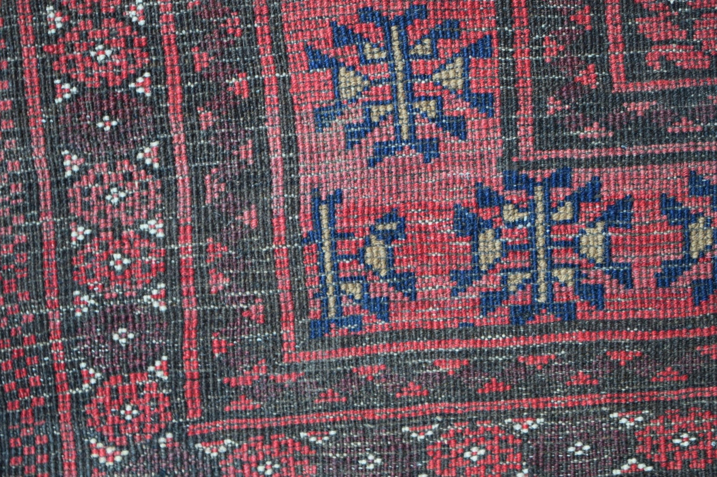 A close-up of a vintage Afghan Baluch rug featuring geometric blue and tan motifs on a red field.