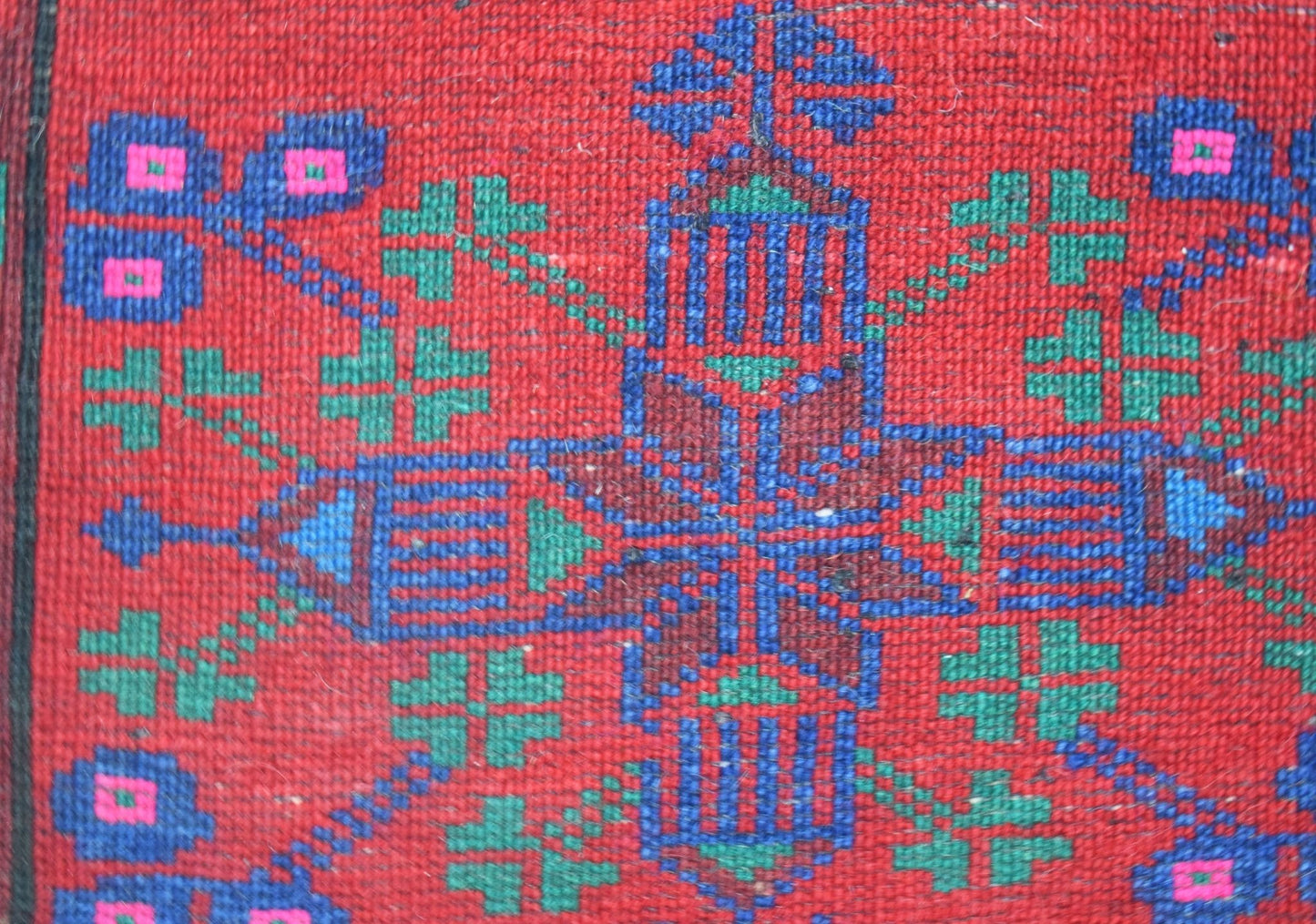A close-up of a vintage handwoven Afghan Baluch rug featuring a geometric floral pattern in red, blue, and green wool.