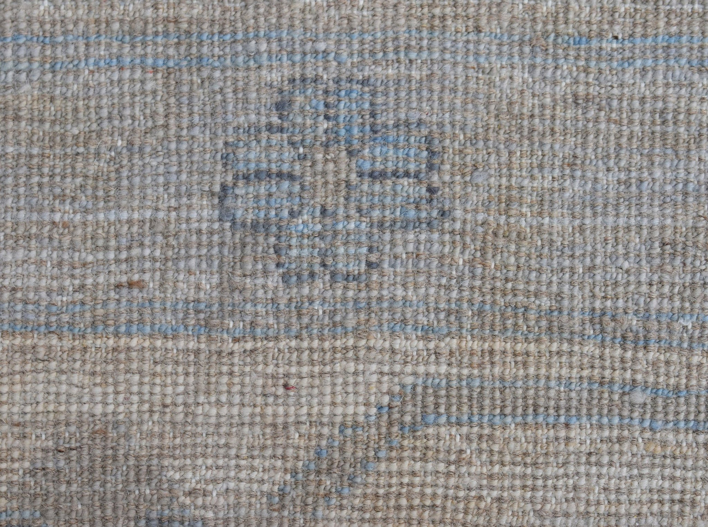 A close-up of a handwoven wool rug featuring a subtle blue floral motif on a neutral beige field.