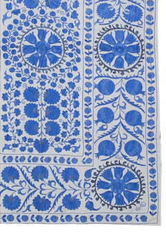 Hand-embroidered Uzbek Suzani featuring blue floral and pomegranate motifs on a white cotton base.