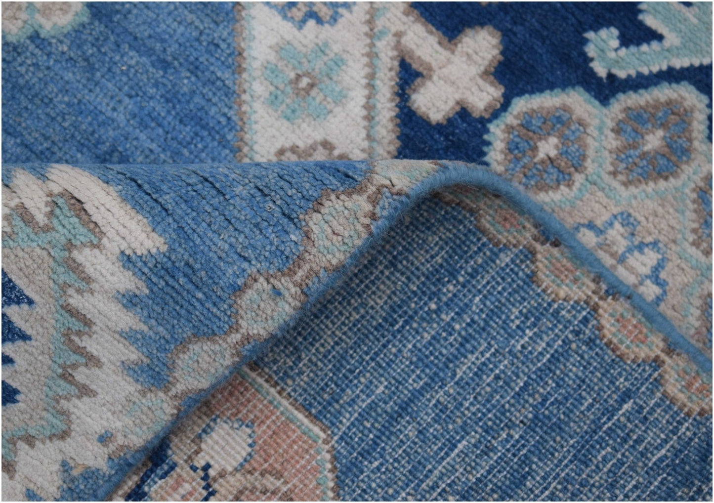 A corner of a blue Afghan Kazakh wool rug is folded back to reveal its woven cotton backing.