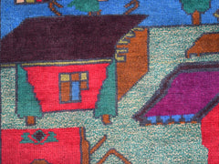 A handmade Afghan wool rug featuring a colorful, stylized village scene with houses and trees.