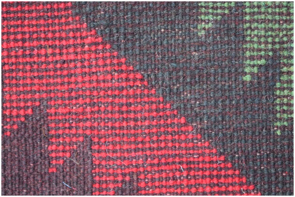 A close-up of a handmade Afghan wool rug featuring geometric patterns in red, black, and green.