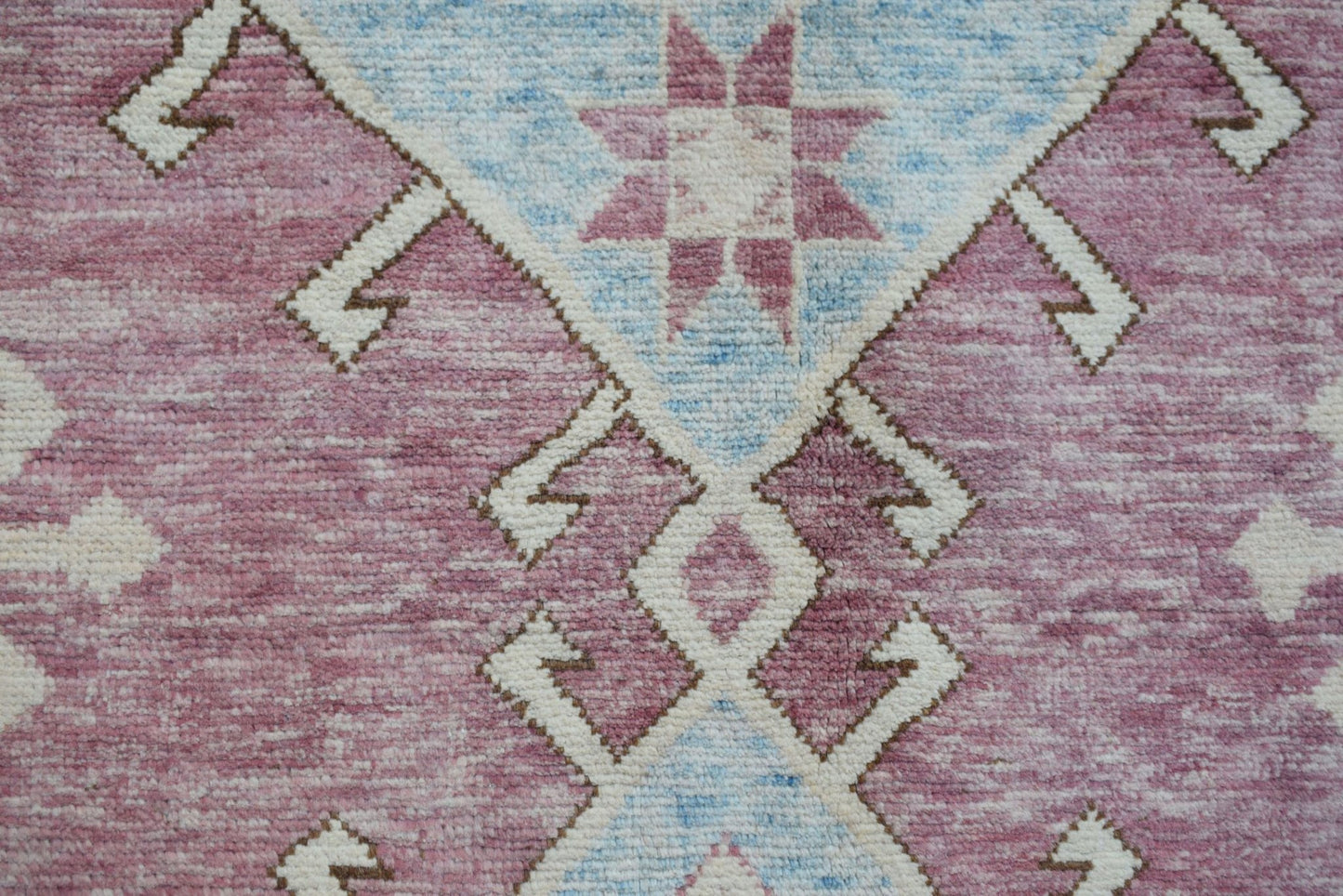 A handmade wool rug featuring a geometric tribal pattern in shades of pink, blue, and cream.