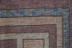 A close-up of a handmade wool rug featuring geometric borders in purple, blue, and cream tones.
