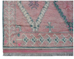 A handmade wool rug featuring a pink background with teal and white geometric tribal patterns.