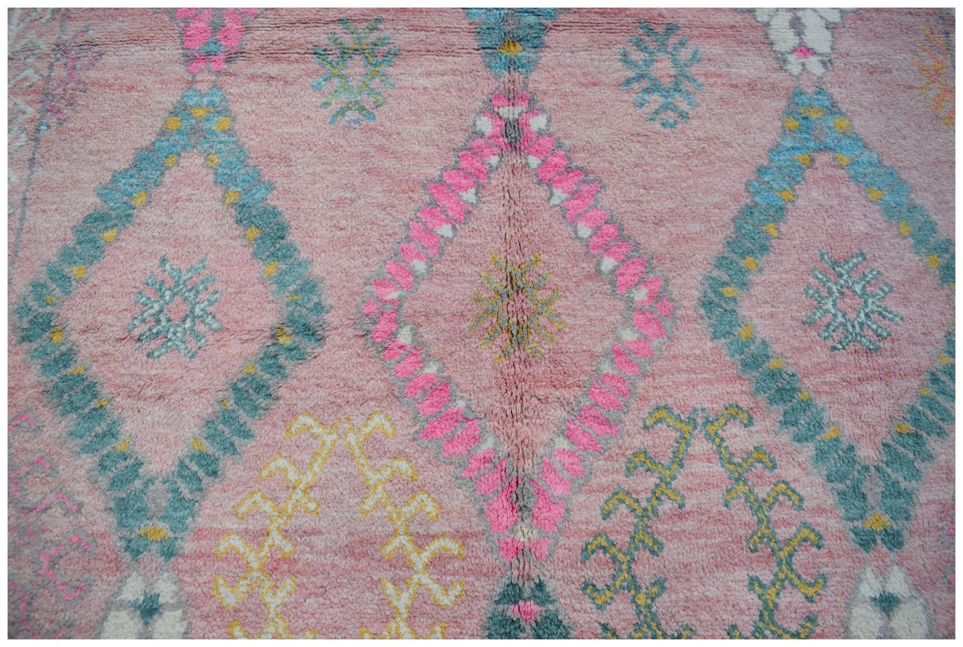 A handmade wool rug featuring a pink background with a repeating diamond pattern in blue and pink.