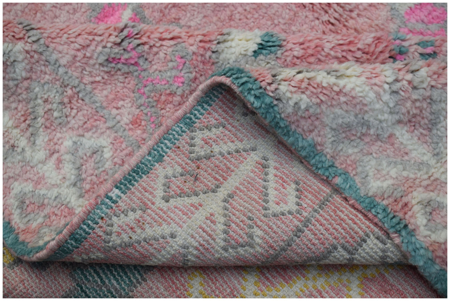 A close-up of a plush, handmade wool rug in shades of pink, white, and teal with a folded corner.