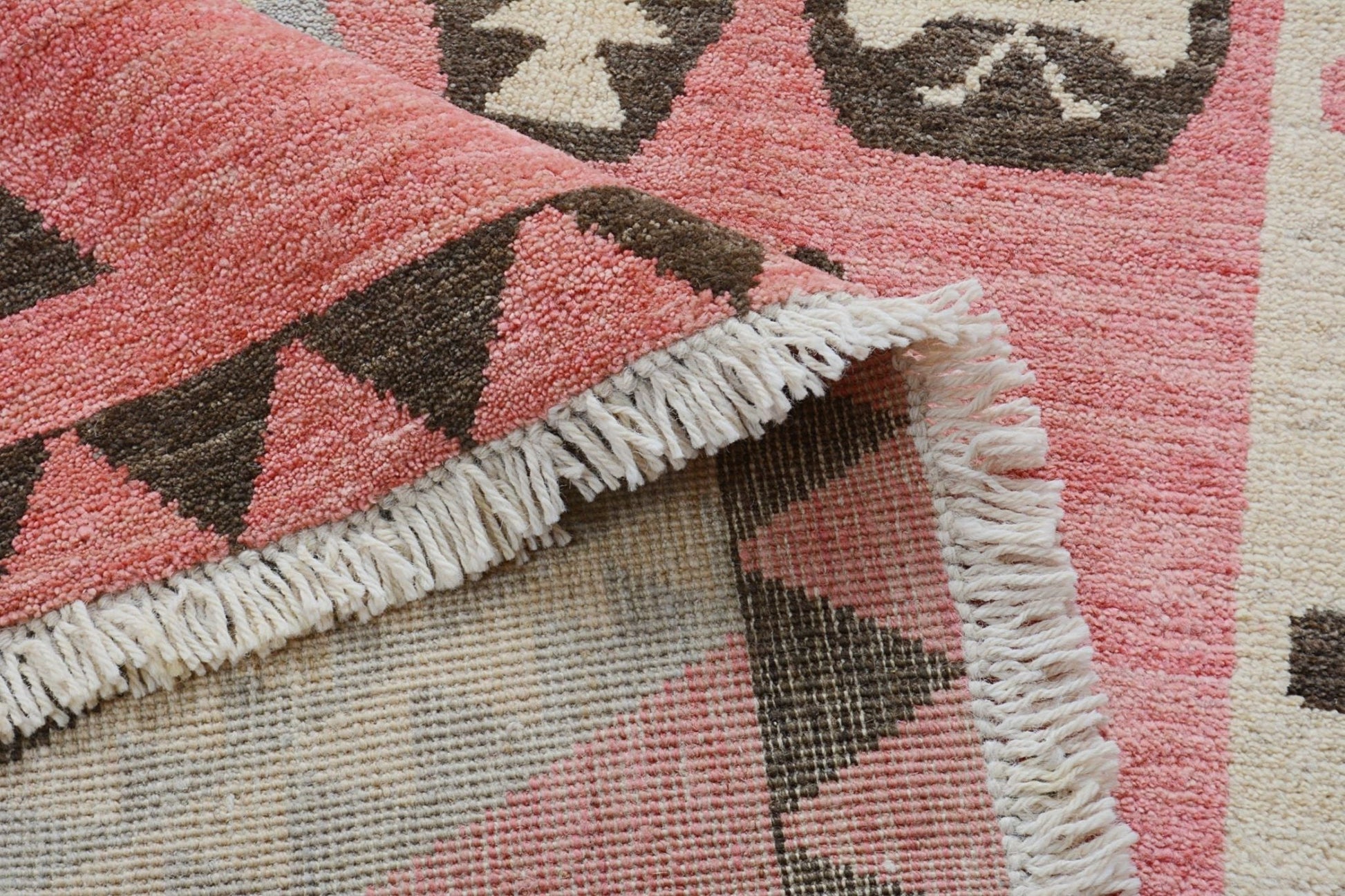 A close-up of a handmade wool rug featuring a pink and brown geometric pattern with fringe edges.
