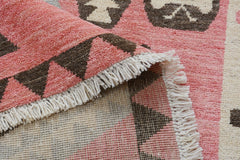 A close-up of a handmade wool rug featuring a pink and brown geometric pattern with fringe edges.