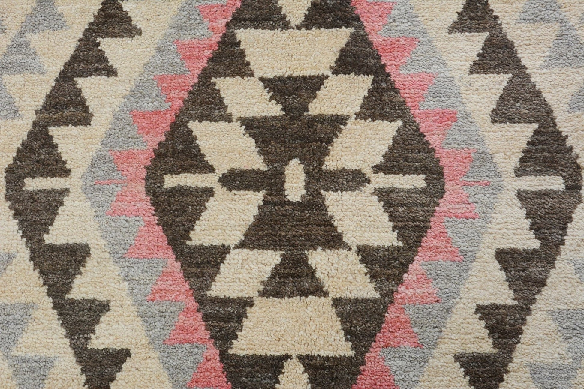 A handmade wool rug featuring a geometric tribal pattern in cream, brown, pink, and grey.