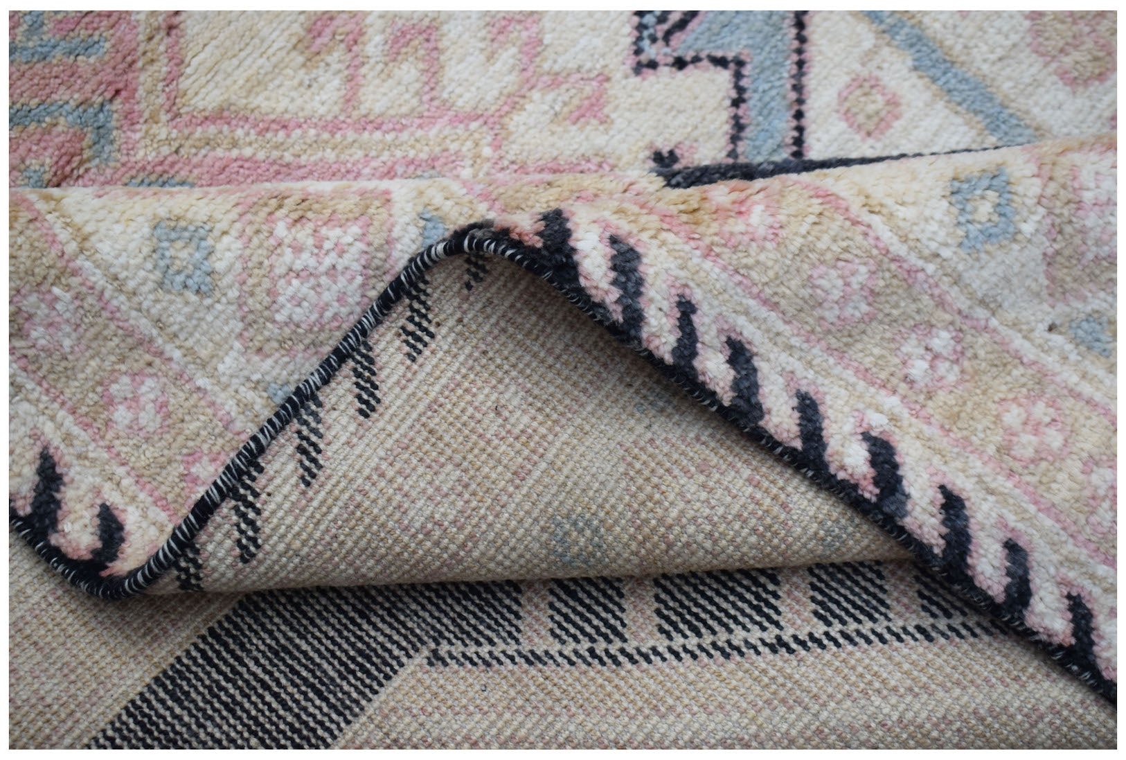 A close-up view of a handmade wool rug featuring a cream, pink, and blue geometric pattern.