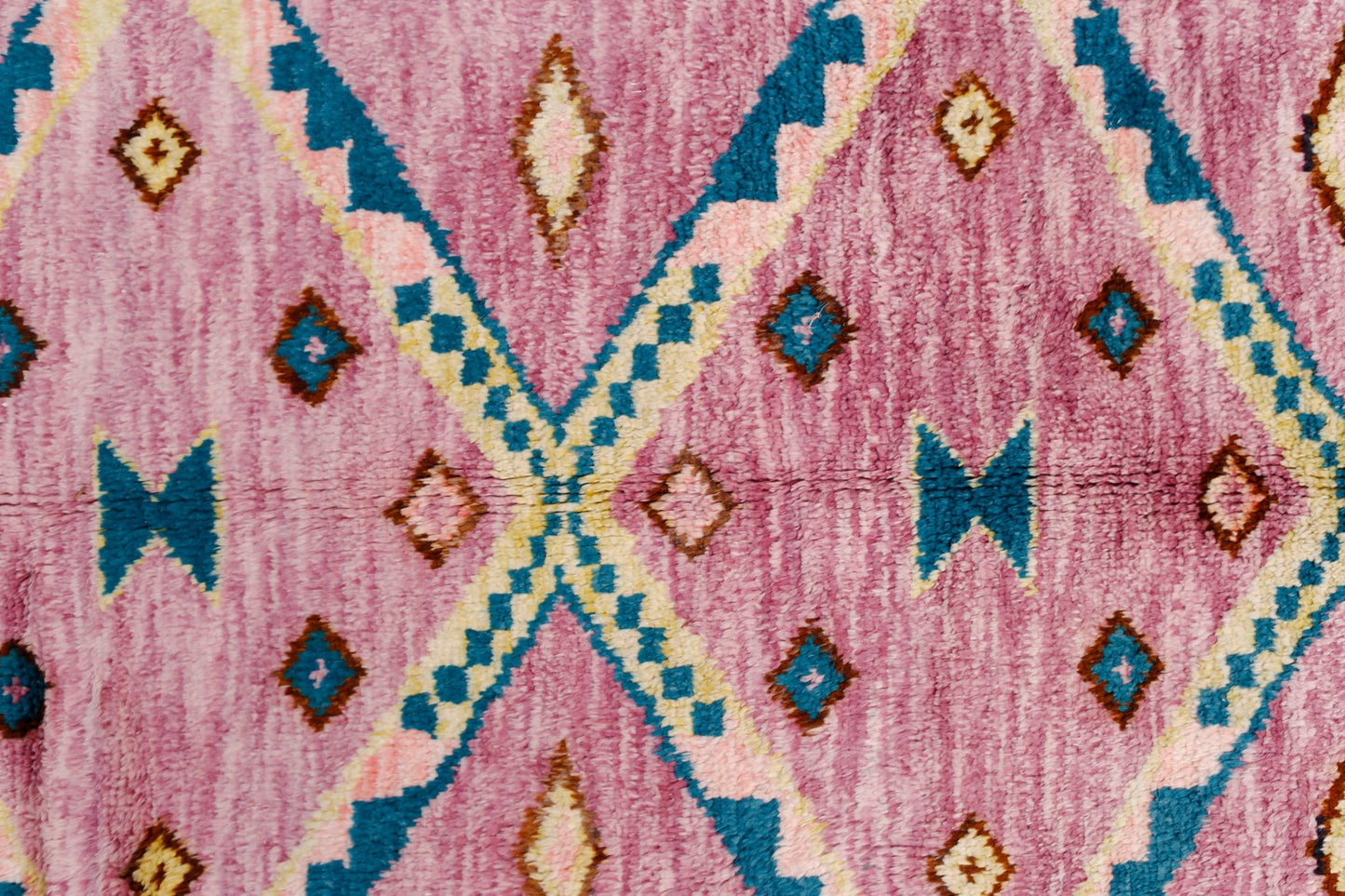 A handmade wool rug featuring a pink background with blue and cream geometric diamond patterns.
