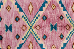 A handmade wool rug featuring a pink background with blue and cream geometric diamond patterns.