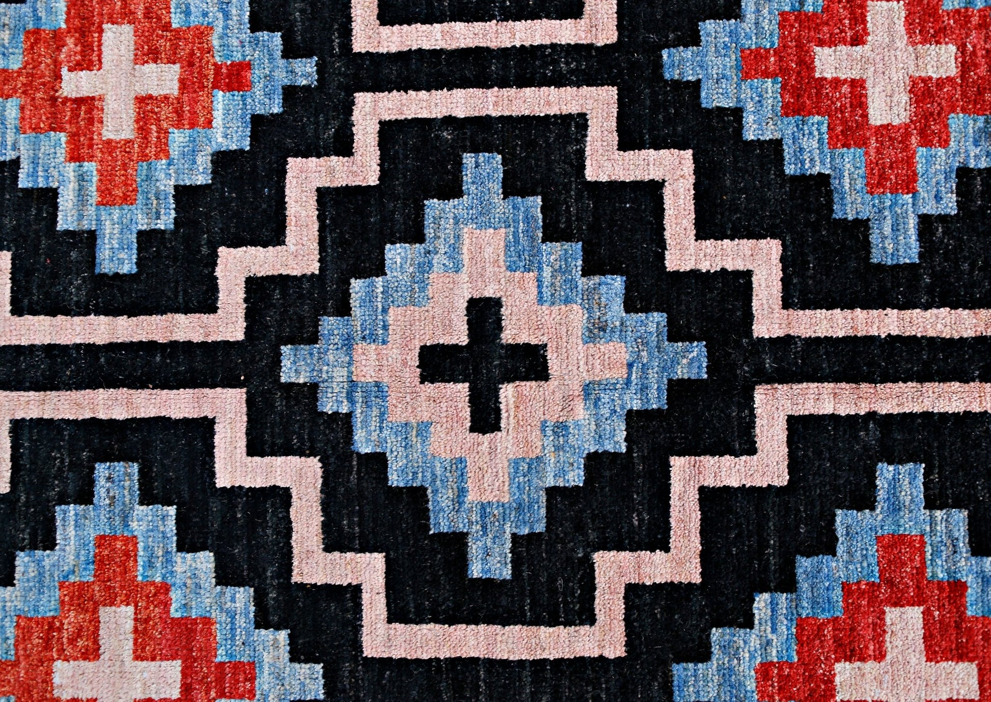 A handmade wool rug featuring a geometric pattern of black, blue, pink, and red diamond motifs.