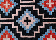 A handmade wool rug featuring a geometric pattern of black, blue, pink, and red diamond motifs.