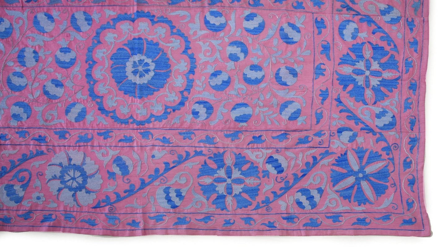 Handmade Uzbek suzani textile featuring blue floral and pomegranate embroidery on a pink background.