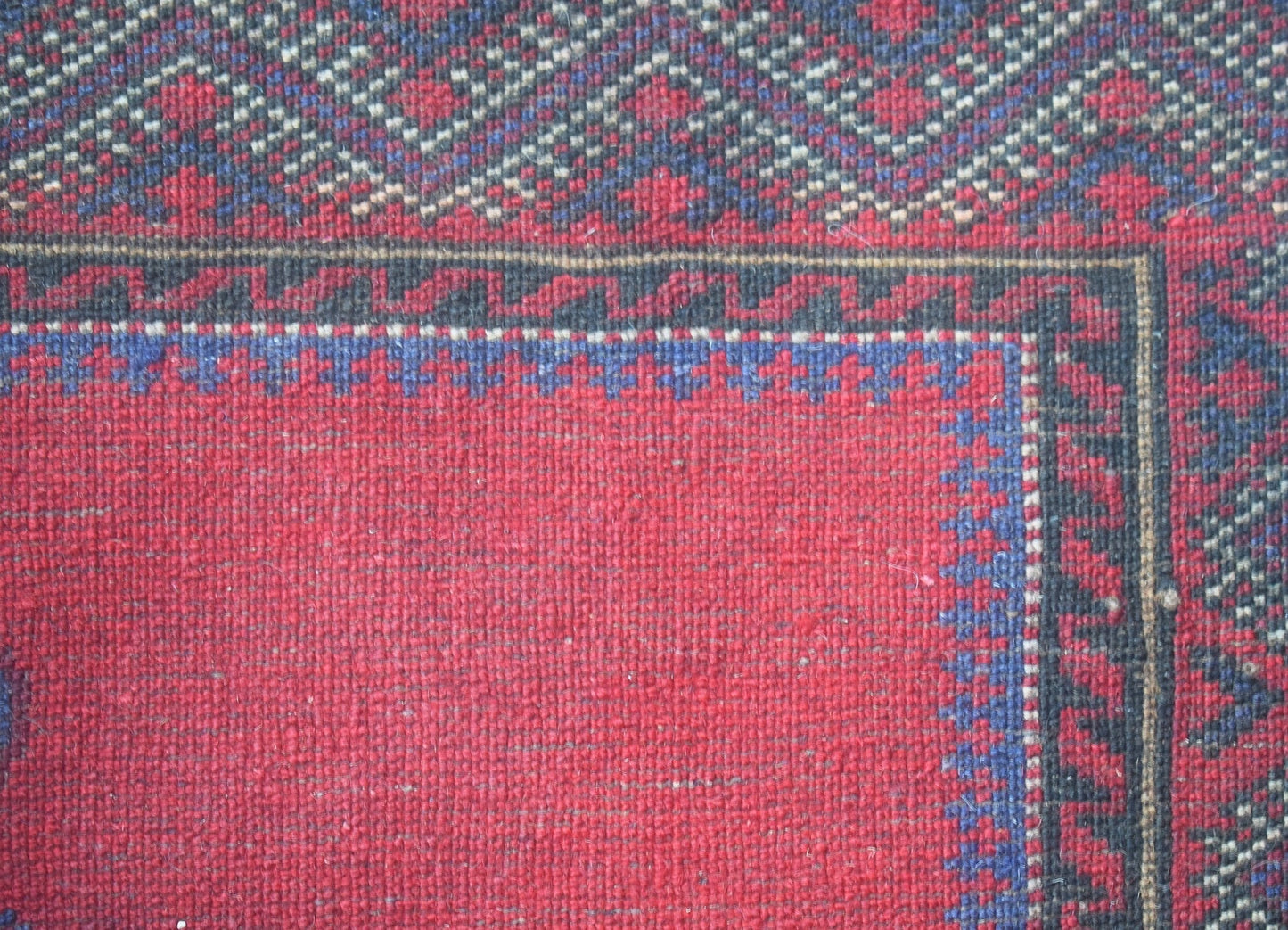 A close-up of a vintage handwoven Afghan Baluch rug featuring a red field and geometric borders.