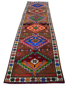 A vintage handmade Kurdish Herki runner featuring colorful geometric tribal motifs on a brown field.