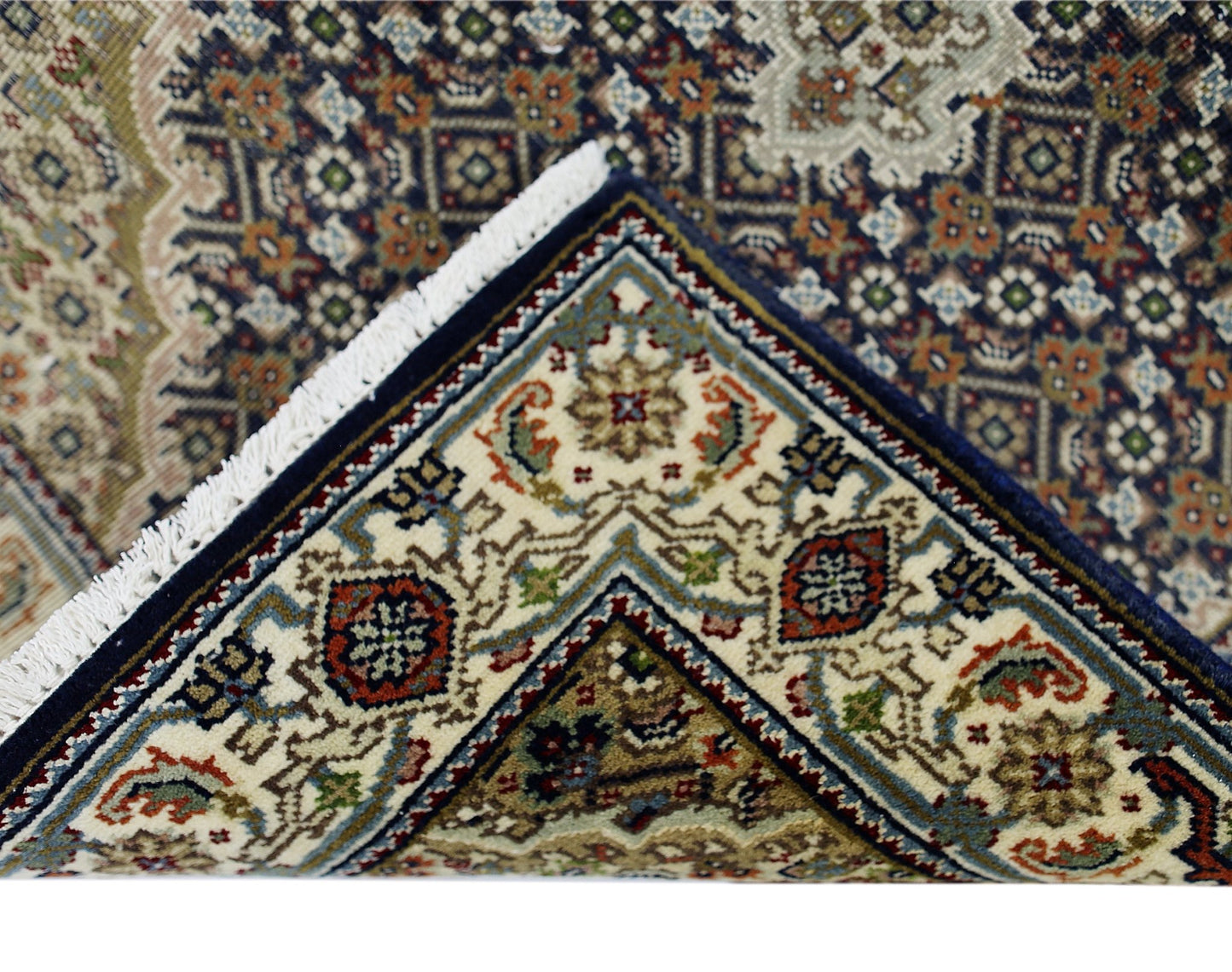 A corner of a vintage Persian silk Tabriz rug with intricate floral patterns and a white fringe.