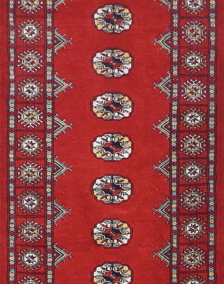 A handmade red wool runner rug featuring repeating geometric Bokhara motifs and patterned borders.