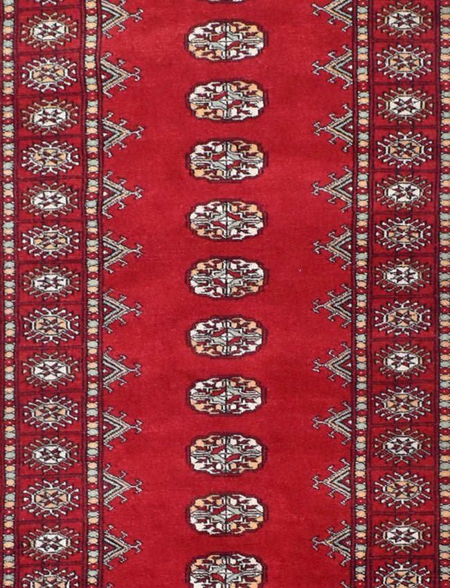 A handmade red wool runner rug featuring repeating geometric Bokhara motifs.