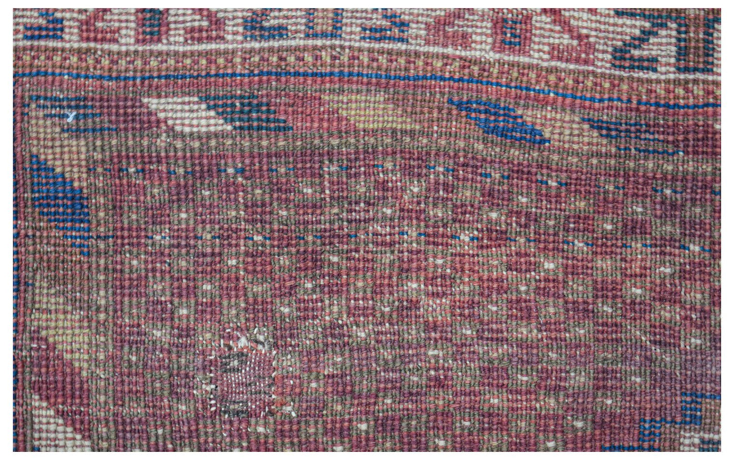 A close-up view of a handwoven antique Persian rug featuring a faded red and blue geometric pattern.