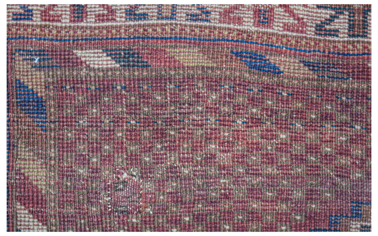 A close-up view of a handwoven antique Persian rug featuring a faded red and blue geometric pattern.