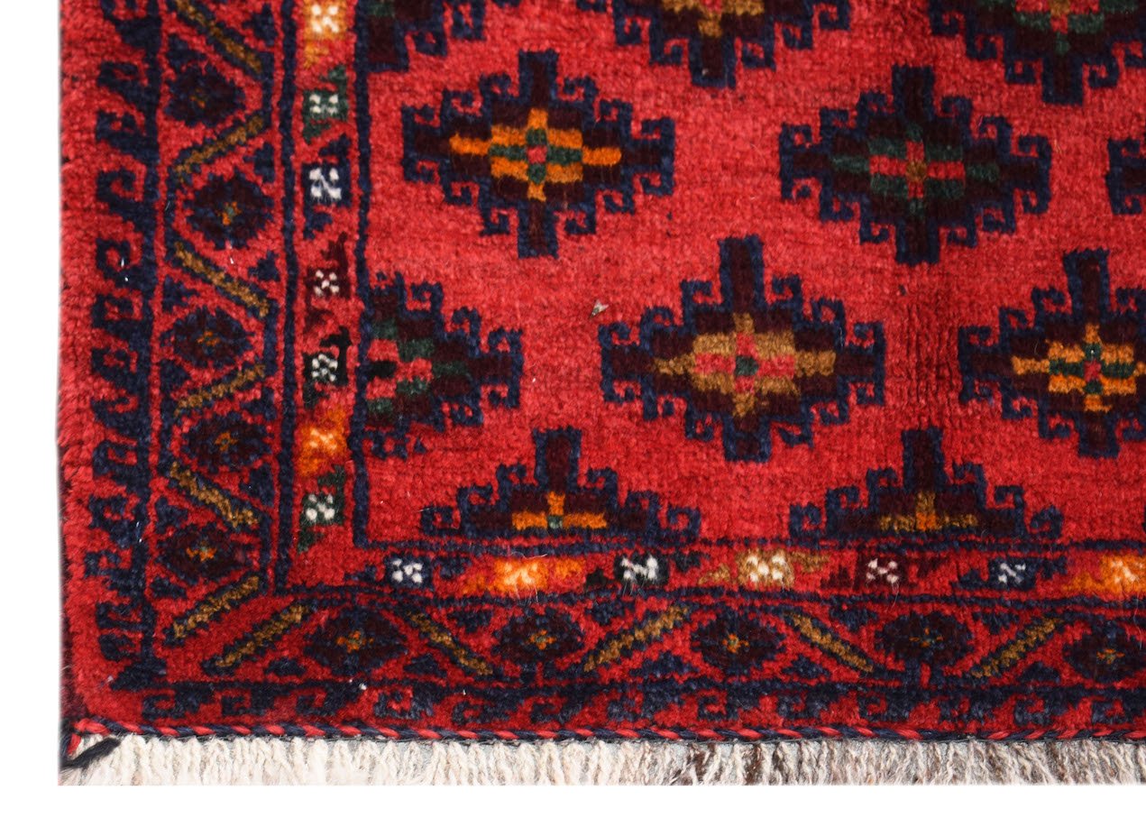 A vintage Afghan Baluch rug featuring a red field with repeating geometric motifs and fringe.