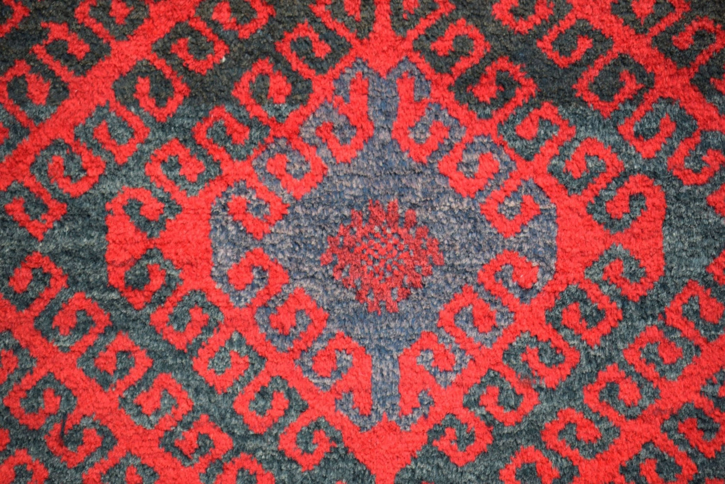 A vintage Afghan Baluch wool rug featuring a repeating red geometric pattern on a dark background.