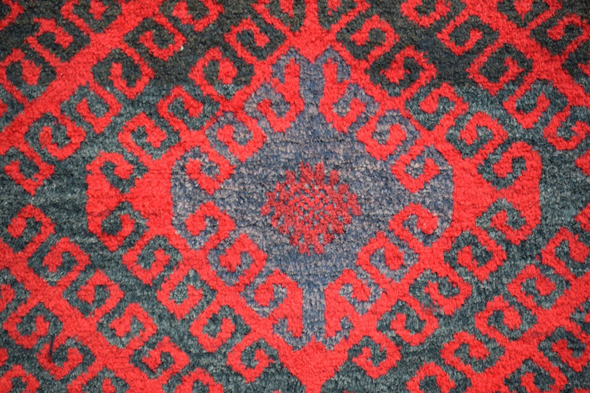 A vintage Afghan Baluch wool rug featuring a repeating red geometric pattern on a dark background.