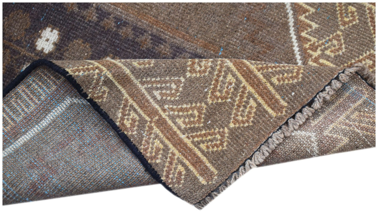 A handmade Afghan Baluch wool rug with a brown geometric pattern and a folded corner.