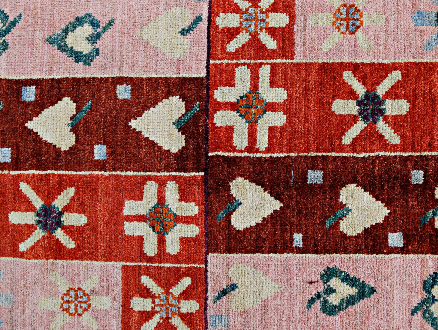 A handmade wool rug featuring a geometric patchwork pattern of floral and leaf motifs in red tones.