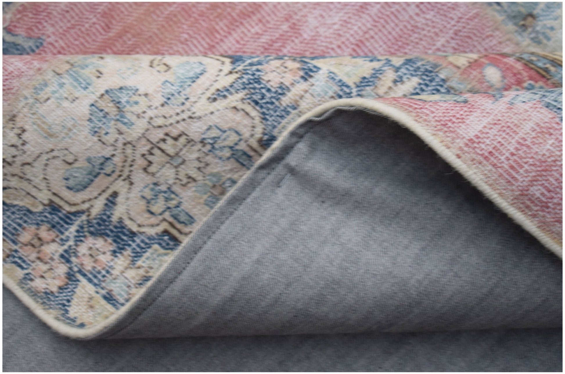 A corner of a vintage patchwork rug with a blue and pink floral pattern and a grey fabric backing.