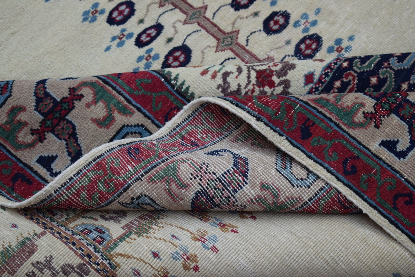 A vintage handwoven Persian rug with a cream field and a folded corner showing a red border.
