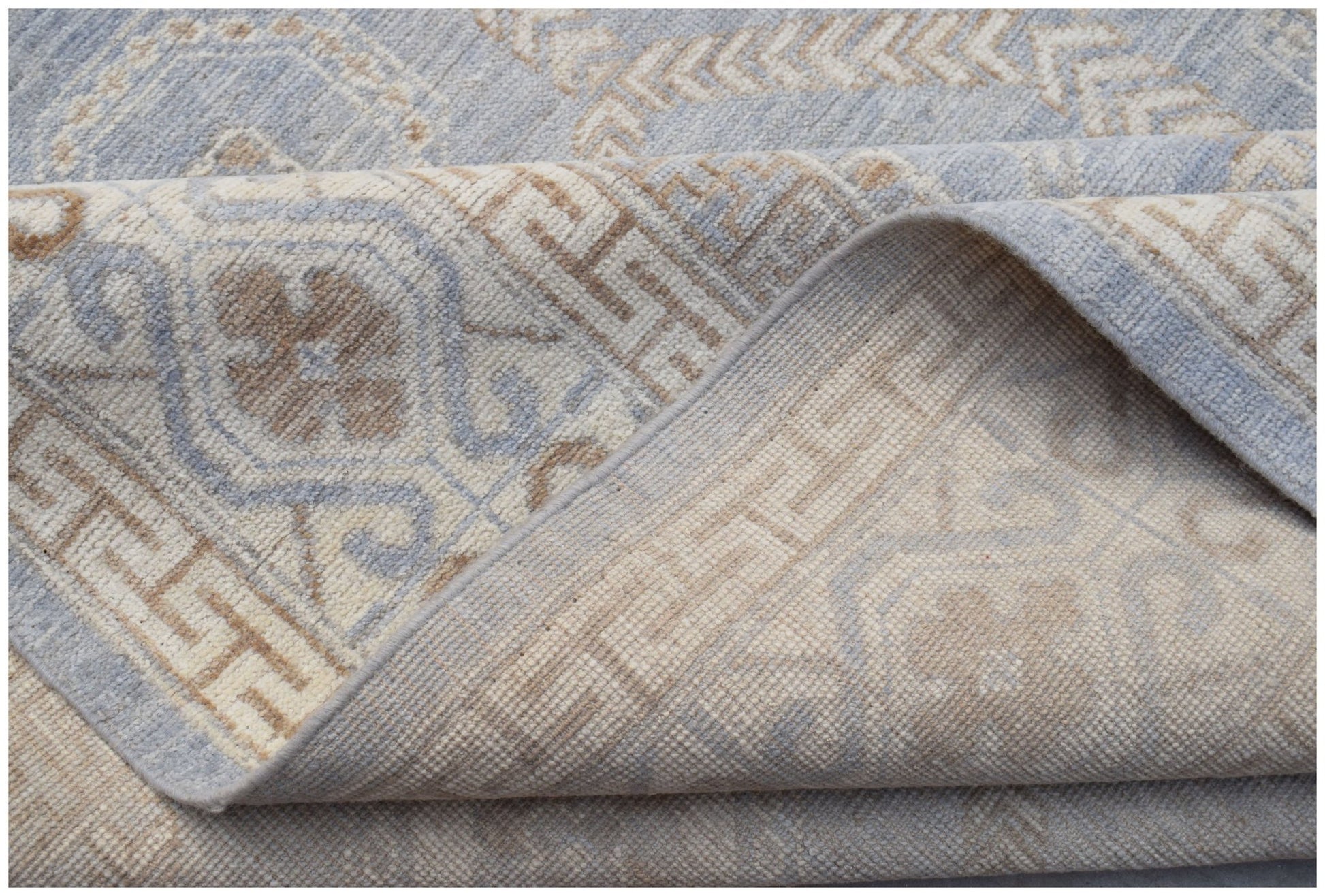 A handmade wool Oushak rug featuring a light blue and beige geometric pattern with a folded corner.