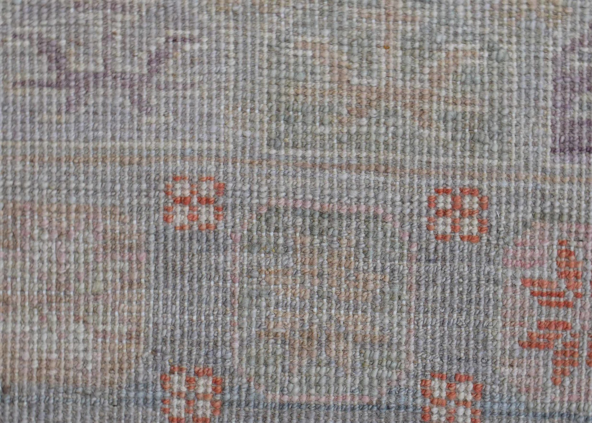 A close-up of a handwoven wool rug featuring a muted, transitional pattern with small orange motifs.