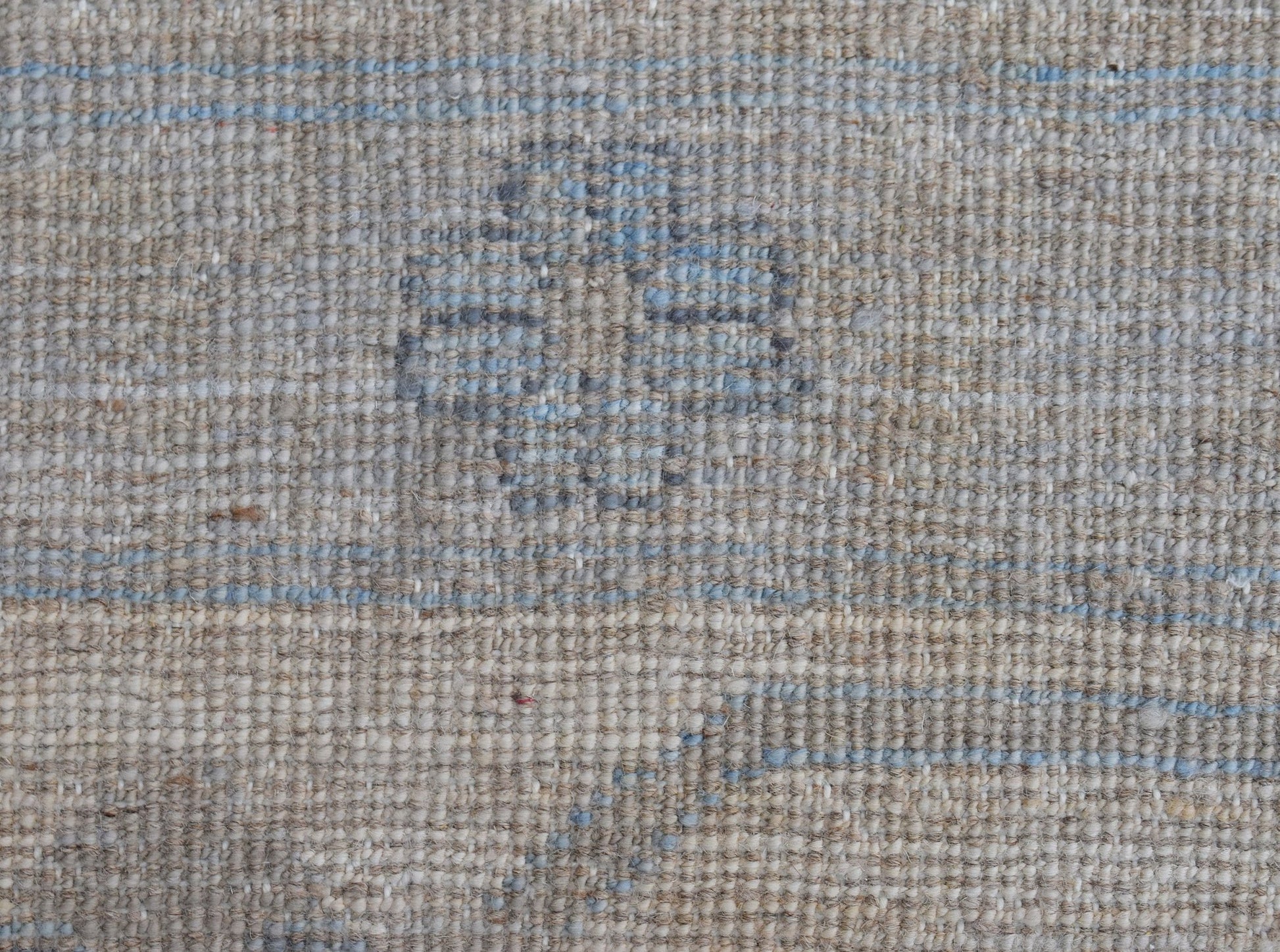 A close-up of a handwoven wool rug featuring a subtle blue floral motif on a neutral beige field.